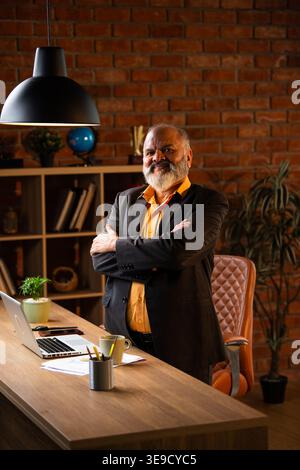 Lächelndes Porträt eines leitenden indischen Geschäftsmannes oder Geschäftsführers, der in einem gemütlichen Büro neben seinem Schreibtisch steht Stockfoto