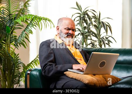 Ein indischer Senior-Geschäftsmann oder Unternehmensleiter arbeitet an seinem Laptop und sitzt bequem auf einem Sofa in einer gemütlichen Büroumgebung mit Profi Stockfoto