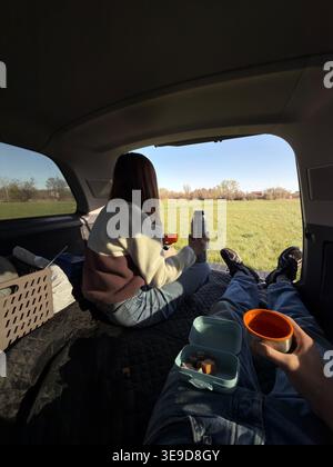Paare entspannen sich im offenen Kofferraum, trinken Kaffee aus der Thermoskanne und Snacks während eines Picknicks auf dem Land. Sie schauen über sonniges Grün gefallen Stockfoto