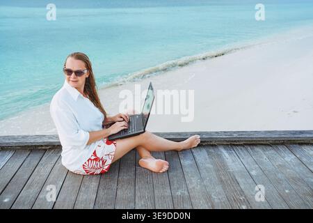 Lächelnde, erfolgreiche Frau mit Laptop an einem schönen, sonnigen Strand. Erfolg. Flexible Fernarbeit, Konzept der Vereinbarkeit von Beruf und Privatleben für Frauen. Stockfoto