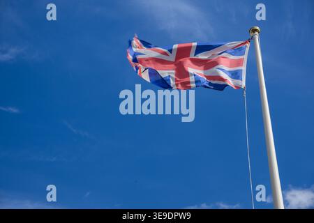 Die Flagge der Union Jack auf einem Fahnenmast vor einem klaren blauen Himmel symbolisiert britische Identität, Patriotismus und Nationalstolz. Stockfoto