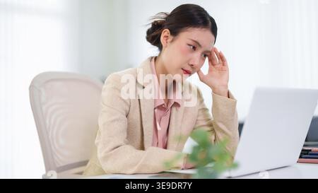 Junge asiatische Geschäftsfrau, die sich bei der Arbeit mit einem Laptop in einem modernen Büro mit Kopfschmerzen gestresst fühlt Stockfoto