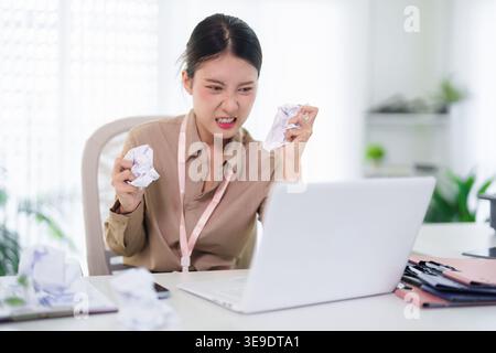 Wütende asiatische Geschäftsfrau zerknittert Papierdokumente frustriert Laptop-Computer, der sich mit der Abgabetermin im Büro beschäftigt Stockfoto