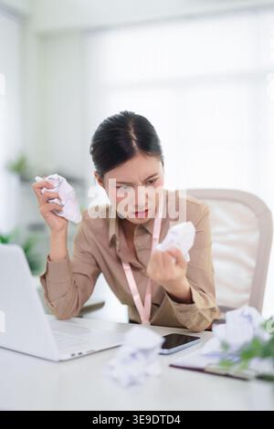Frustrierte asiatische Geschäftsfrau, die mit Arbeitsstress zu kämpfen hat und Papier am Bürotisch zerknittert, die Burnout und Deadline zeigen Stockfoto