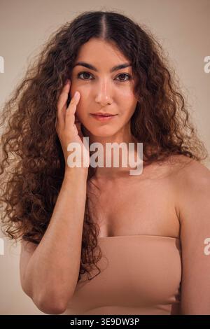 Eine Frau mit lockigem Haar berührt sanft ihr Gesicht und strahlt Selbstvertrauen und Schönheit in einer neutralen Umgebung aus, die ihre natürliche Eleganz und Ausgewogenheit unterstreicht Stockfoto