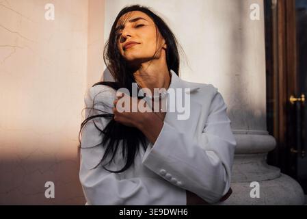 Stilvolle Frau in einem weißen Blazer posiert vor einer strukturierten Wand, hält einen Moment der Selbstsicherheit und Eleganz fest und unterstreicht zeitgenössische Mode und persönlichen Stil Stockfoto