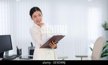 Asiatische Geschäftsfrau mit Klemmbrett, die in hellen modernen Büroarbeitsplätzen steht und selbstbewusst professionell lächelt Stockfoto