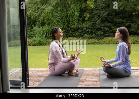 Verschiedene weibliche Freunde in Yoga-Kleidung sitzen auf Yoga-Matten auf der Terrasse und meditieren mit Mudra. Outdoor, Achtsamkeit, Gelassenheit, Lifestyle, Wellness, Terrasse, Grün Stockfoto
