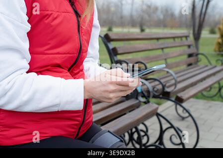 Frau mit Sportkleidung und Kniebeuge oder Orthese nach Beinoperationen im Park mit Smartphone. Medizin- und Gesundheitskonzept. Stockfoto