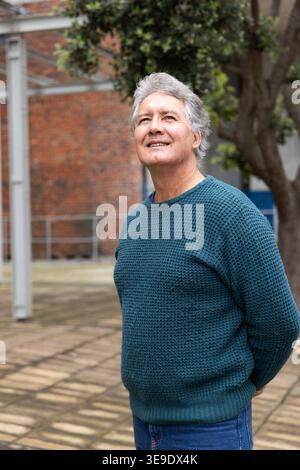Ein älterer Mann steht im gepflasterten Innenhof, trägt einen blauen Strickpullover und eine blaue Jeans und blickt nach oben. Älter, Gentleman, Outdoor, Urban, Ziegelstein, Baum, Tür Stockfoto