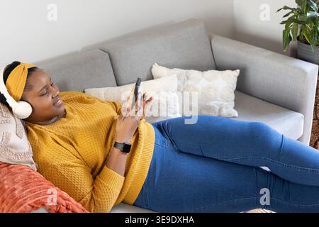 Afroamerikanerin, die zu Hause auf einem grauen Sofa liegt, Senfpullover trägt und Smartphone hält. Gelb, Strick, Couch, Kissen, Telefon, Kopfhörer, gemütlich Stockfoto