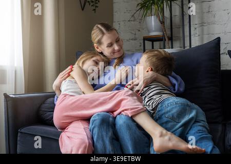 Mutter mit zwei Kindern, die sich im Wohnzimmer auf einer gemütlichen Couch umarmen. Mutter mit liebenswerter, liebevoller Tochter und Sohn, die Zeit miteinander verbringen. Familienbindung, tr Stockfoto