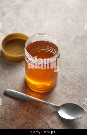 Offenes Glas mit flüssigem Honig auf strukturierter grauer Steinoberfläche mit Metalllöffel und goldenem Deckel, Blick von oben mit Kopierraum, kulinarisches Konzept für Bio-Lebensmittel Stockfoto