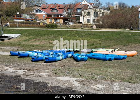 Deutschland, Mecklenburg Vorpommern, Rerik, 8. April 2026: Bunte Kajaks ruhen auf dem grasbewachsenen Strand in Ufernähe mit Ferienhäusern im Hinterland Stockfoto