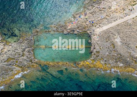 Blick aus der Vogelperspektive auf den Qawra Pool, ein rechteckiger, in Kalkstein geschnittener Pool neben klarem türkisfarbenem Wasser an der maltas felsigen Küste in St Pauls Bay. Stockfoto