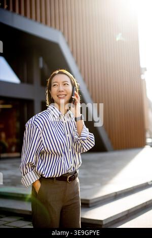 Eine Koreanerin steht vor einem modernen Gebäude und spricht mit ihrem Telefon. Sie lächelt und trägt ein Hemd mit Streifen und grauer Hose. Stockfoto