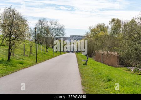 Ein gepflasterter Weg führt durch eine grüne Parklandschaft mit Bäumen und Bänken. Die Szene zeigt eine friedliche Umgebung im Frühling. Stockfoto