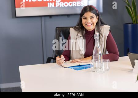 Frau, die am Konferenztisch sitzt und den Stift über offenem Notizbuch mit blauem Ordner hält Stockfoto