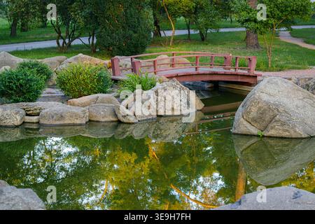 Gartenszene mit einer kleinen roten Brücke, die sich über einen Teich erhebt, umgeben von großen Felsen, üppigem Grün und Bäumen, die sich im Wasser spiegeln Stockfoto
