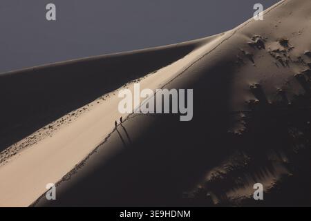 Zwei Personen spazieren entlang eines Sanddünen-Berges mit Fußspuren und langen Schatten in der Namib-Wüste, Namib-Naukluft Park, Deadvlei, Namibia. Stockfoto