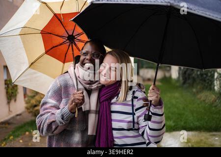 Verschiedene Freundinnen lächelnd und halten Regenschirme, die im Regen stehen Stockfoto