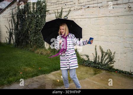 Fröhliche junge Frau, die im Regen tanzt und Schirm und Telefon hält Stockfoto