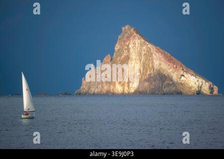 Ein kleines Boot segelt am vulkanischen Meereshaufen von Strombolicchio vorbei, während sich über der Insel Stromboli, Italien, Sturmwolken sammeln. Die Insel war der Vulkandübel, der während einer massiven Explosion vor 200.000 Jahren aus dem Stromboli-Vulkan geworfen wurde. Stockfoto