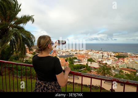 Frau, die mit ihrem Smartphone ein Foto von der Panoramablick auf die Stadt und das Meer von einem Aussichtspunkt in Puerto de la Cruz, Teneriffa, Kanarischen Inseln macht. Stockfoto