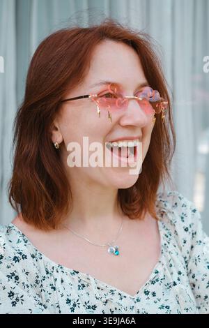 Fröhliche Frau mit dunkelbraunem Haar lacht, während sie eine einzigartige rosa Wolkenbrille trägt. Freude, Positivität und sorgloser Geist. Ideal für Lifestyle oder Stockfoto