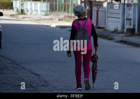 Eine Person läuft entlang einer Asphaltstraße, trägt einen Rucksack und einen geschlossenen Schirm bei Tageslicht. Stockfoto