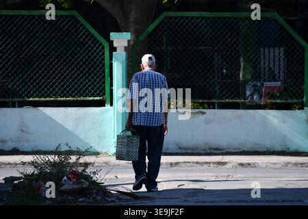 Ein älterer Mann in einem karierten Hemd, der eine Korbtasche trägt, während er auf einer städtischen Straße an einem verwitterten grünen Zaun vorbeiläuft Stockfoto