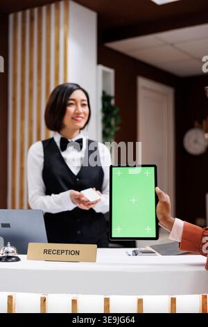 Frau, die an der Rezeption ein Tablett mit grünem Bildschirm hält, erhält eine Karte für den Zimmerzugang von der asiatischen Rezeptionistin. Junge Person, die an der Hotelrezeption ein isoliertes Modell mit Chromakey benutzt. Stockfoto