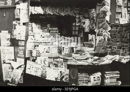Historisches Foto mit dem Titel Woman Reading Inside Zeitungskiosk, das einen überfüllten Kiosk mit Zeitungen, Zeitschriften und gedruckten Medien zeigt, während eine Frau im Kiosk liest. Foto von Angelo Rizzuto, November 1953 Stockfoto