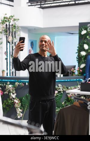 Shopping Mall Boutique arabischer Mann Kunde winkt hallo, während er Selfie auf dem Smartphone macht. Ein junger lächelnder Blogger begrüßt Social-Media-Follower, während er im Bekleidungsgeschäft Live-Übertragungen ausstrahlt Stockfoto