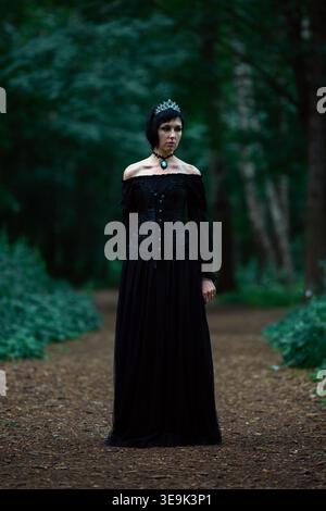 Junge Frau, gekleidet als dunkle gotische Königin, die auf einem Waldweg steht, trägt ein langes schwarzes Korsett-Kleid, eine blaue Edelsteinkrone und einen Chok Stockfoto