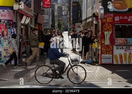Ein älterer Japaner, der eine chirurgische Maske gegen COVID-19 trägt, fährt mit dem Fahrrad an einer belebten Straße in Shin Okubo, Tokio, vorbei. Stockfoto