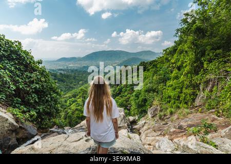 Junge Frau, die auf einer felsigen Klippe mit Blick auf das üppige Dschungeltal und die fernen Hügel in der Nähe des Samui Wasserfalls in Thailand steht Stockfoto