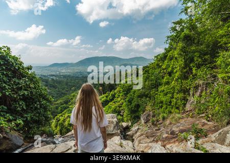 Junge Frau, die auf einem felsigen Felsvorsprung mit Blick auf das üppige tropische Tal und die fernen Berge in der Nähe des Samui Wasserfalls in Thailand steht Stockfoto