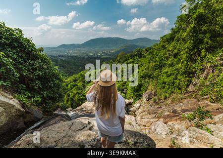 Eine junge Frau mit Strohhut auf einem felsigen Felsvorsprung mit Blick auf das üppige tropische Tal und die fernen Berge in der Nähe des Samui Wasserfalls in Thailand Stockfoto
