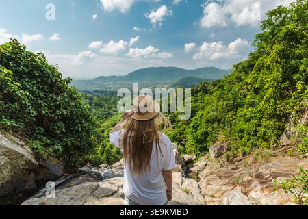 Junge Frau mit Strohhut, die auf dem felsigen Aussichtspunkt des Wasserfalls mit Blick auf das üppige grüne Dschungeltal und die fernen Berge auf der Insel Samui steht Stockfoto