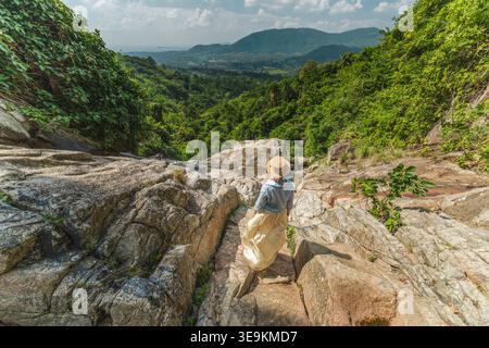 Junge Frau mit Strohhut, die am felsigen Wasserfallrand mit Blick auf das üppige grüne Tal und die Berge auf Koh Samui Thailand steht Stockfoto
