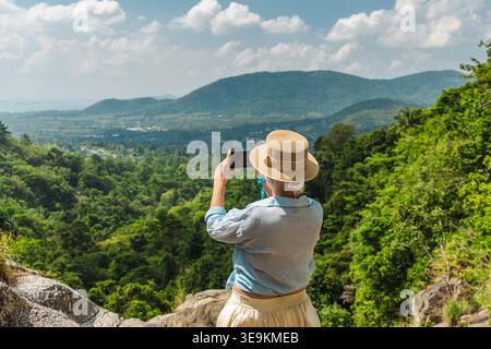 Junge Frau mit Strohhut, die Fotos mit dem Smartphone am tropischen Aussichtspunkt in den Bergen, dem üppigen grünen Tal, macht Stockfoto