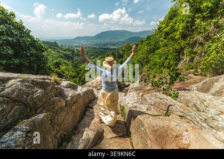 Junge Frau, die auf felsigem Aussichtspunkt mit erhobenen Armen mit Blick auf das üppige tropische Tal und ferne Hügel steht, Freiheit und Abenteuer Konzept Stockfoto