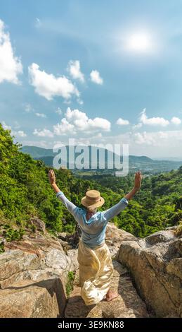 Junge Frau, die auf einem felsigen Aussichtspunkt mit erhobenen Armen mit Blick auf das üppige tropische Tal und entfernte Hügel steht Stockfoto