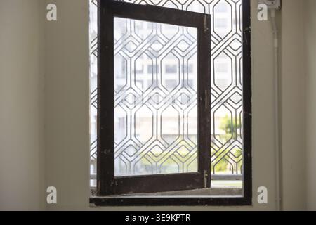 Blick durch ein offenes Fenster mit dunklem Rahmen und geometrischem Metallgitter und ruhiger Innenraum, der von weichem Tageslicht beleuchtet wird Stockfoto