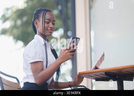 Junge schwarze Geschäftsfrau, die ihr Smartphone neben einem Tablet an einem Café-Tisch im Freien benutzt. Mobile Kommunikation, Multitasking, Online-Workflow, Remote-Arbeit Stockfoto