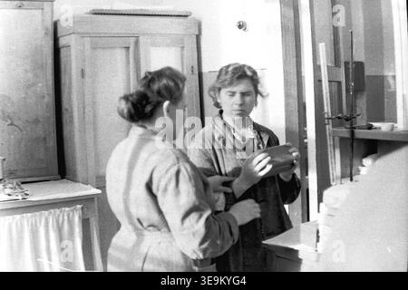 Slawjansk, 1950er Jahre AIZ-Werk. Ein wunderschönes historisches Schwarzweiß-Archivfoto, das zwei sowjetische Arbeiter in einer Produktionsanlage zeigt. Im Vordergrund steht die Arbeiterin Valentina Timofeevna Kravelis, die spezielle technische Ausrüstung hält, die die Frauen gemeinsam sorgfältig überprüfen. Dieses authentische Bild vermittelt lebendig Momente der Interaktion am Arbeitsplatz, typische Industriekleidung und aufrichtige zwischenmenschliche Kommunikation in einem großen sowjetischen Unternehmen und demonstriert den Alltag und den friedlichen Donbass. Stockfoto