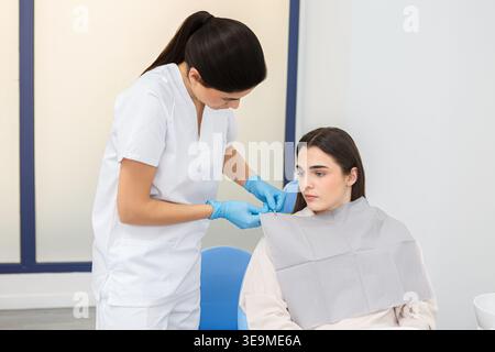 Ein Dentalhygieniker legt einem Patienten in einer Zahnklinik ein Lätzchen an. Der Patient sitzt auf dem Stuhl; der Termin ist für eine routinemäßige Delle Stockfoto