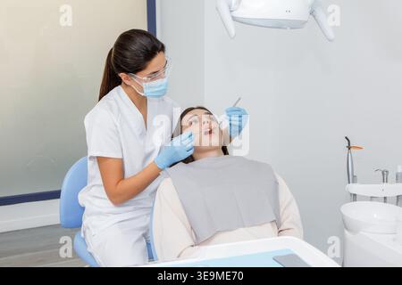 Ein Zahnarzt im weißen Mantel untersucht einen Patienten, der auf einem Zahnarztstuhl sitzt. Der Patient trägt ein Lätzchen, während der Zahnarzt Zahnwerkzeuge zur Untersuchung verwendet. Stockfoto