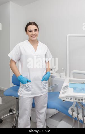 Ein Zahnarzt steht in einer Zahnklinik lächelnd und trägt eine weiße Uniform mit blauen Handschuhen. Die Klinik ist modern und gut beleuchtet und mit Zahngeräten ausgestattet Stockfoto